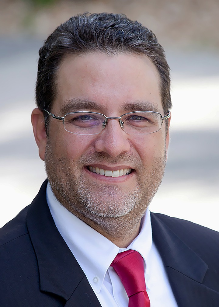 Sam Kaufman in a dark suit and red tie over a blurred background