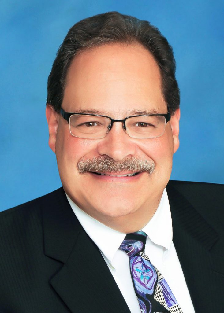 Greg Ross in a dark suit and colored tie over a blue background